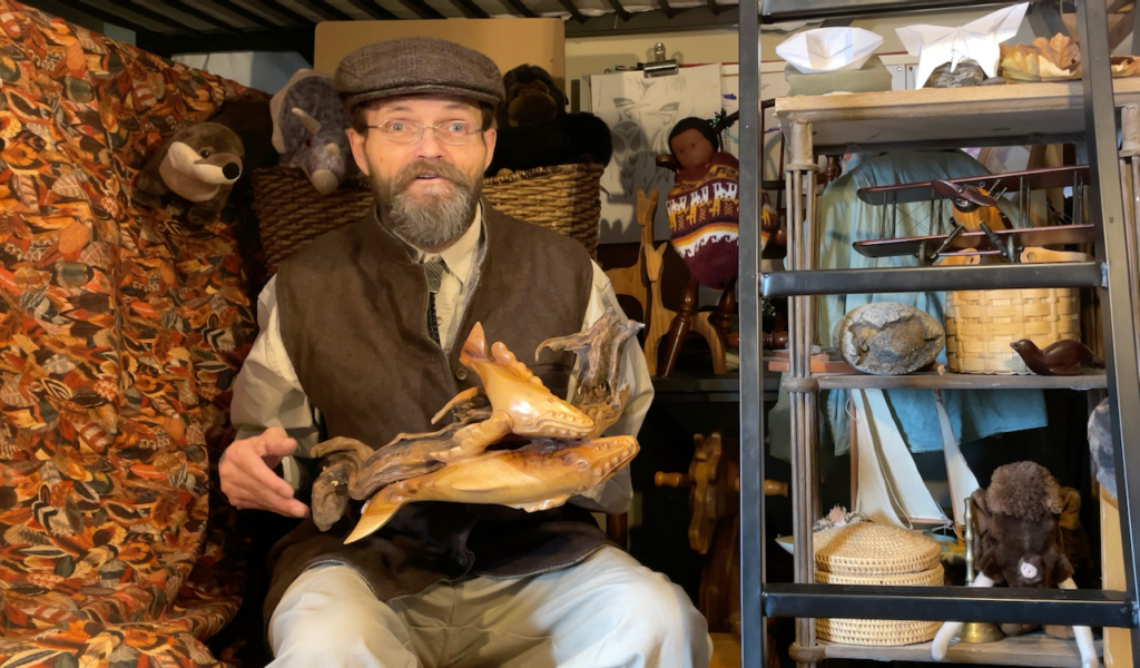 Laird Fetzer Hamblin sitting, singing a story, surrounded by toys and holding a unique piece of driftwood upon which a wooden carving of a mother and infant whale is attached.
